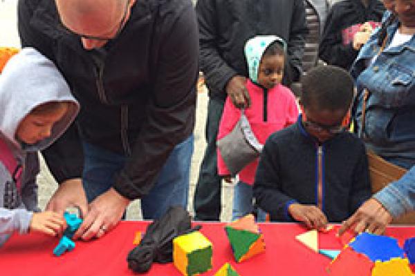 Children and adults playing with mathematical objects (soma cube, tangram, platonic solids) at an exhibit.
